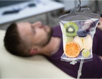 Patient auf einer Liege mit einer Infusion, die mit Obst wie Orange, Kiwi und Trauben gefüllt ist, symbolisch für Vitamintherapie